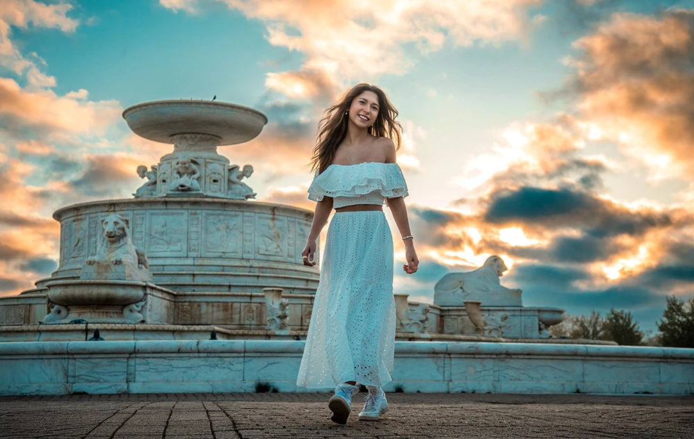 A high school senior girl in a white off-shoulder top and long skirt walks and smiles in front of a large ornate fountain at sunset, with dramatic clouds and warm sunlight in the sky behind her.
