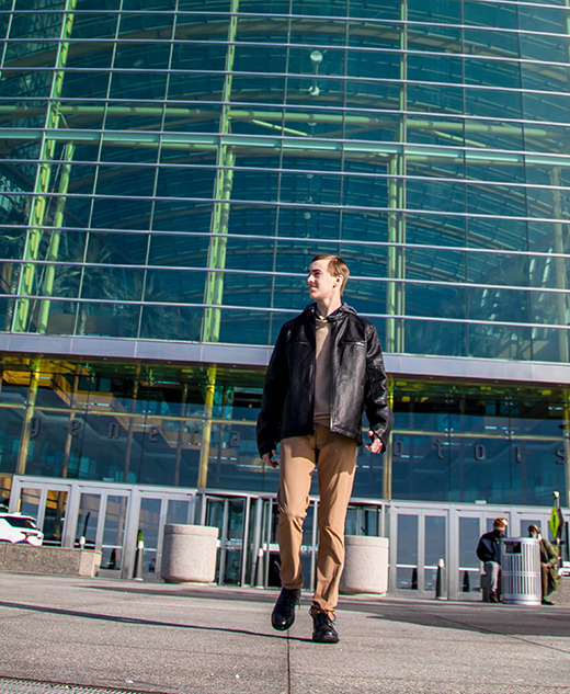 A high school senior guy in a black jacket and tan pants walks outside a modern glass building, with cars and people in the background on a sunny day—perfect for capturing stylish urban senior portraits.