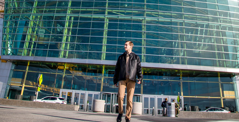 A high school senior guy in a black jacket and tan pants walks outside a modern glass building, with cars and people in the background on a sunny day—perfect for capturing stylish urban senior portraits.