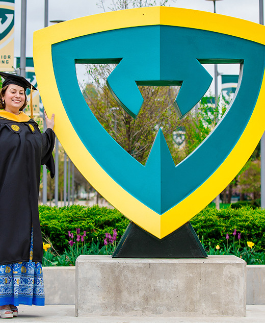 A graduate in a cap and gown stands smiling next to a large Wayne State University shield sculpture, with campus buildings, green foliage, and Warrior Strong banners in the background.