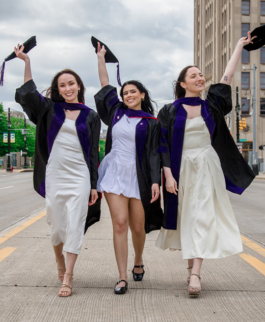 Three women in graduation gowns smile and walk down an empty city street, holding their caps in the air. Dressed in white and light blue, they celebrate with law school graduation pictures as tall buildings and trees frame the background.