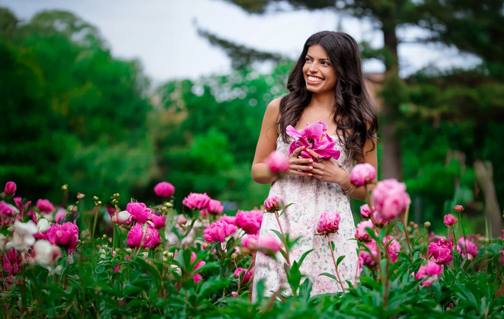 A high school senior girl in a floral dress stands in a garden at Nichols Arboretum, surrounded by blooming pink peonies, holding a bouquet and smiling, with green trees in the background—a perfect senior pictures session.
