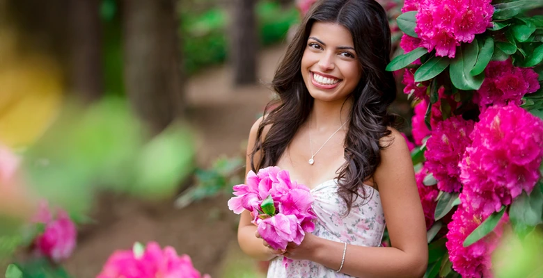 A high school senior girl with long dark hair and a floral dress smiles while holding pink flowers, standing beside a bush full of bright pink blossoms during her Nichols Arboretum senior pictures session in a lush garden setting.