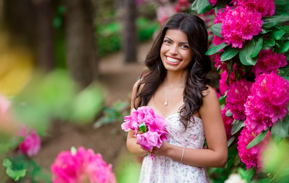 A high school senior girl with long dark hair and a floral dress smiles while holding pink flowers, standing beside a bush full of bright pink blossoms during her Nichols Arboretum senior pictures session in a lush garden setting.