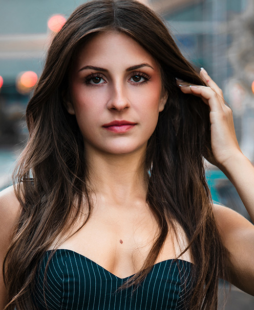 A high school senior girl with long brown hair in a strapless, dark dress stands outside on a city street near a Porsche, confidently looking at the camera. The background is softly blurred, perfect for Northville senior pictures in downtown Detroit.