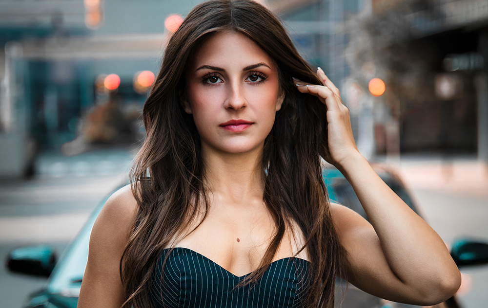 A high school senior girl with long brown hair in a strapless, dark dress stands outside on a city street near a Porsche, confidently looking at the camera. The background is softly blurred, perfect for Northville senior pictures in downtown Detroit.