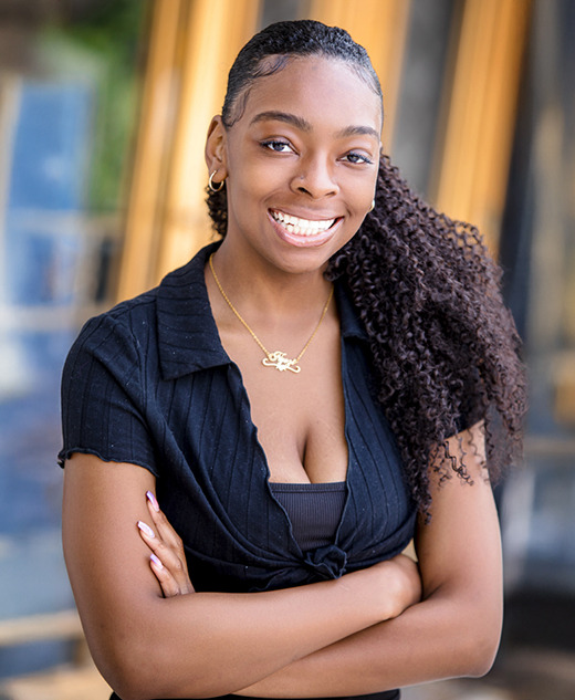 A high school senior girl with long curly hair, wearing a black top and gold necklace, stands outdoors with her arms crossed, smiling at the camera during her River Rouge senior pictures session. The background features blurred blue and yellow architectural elements.