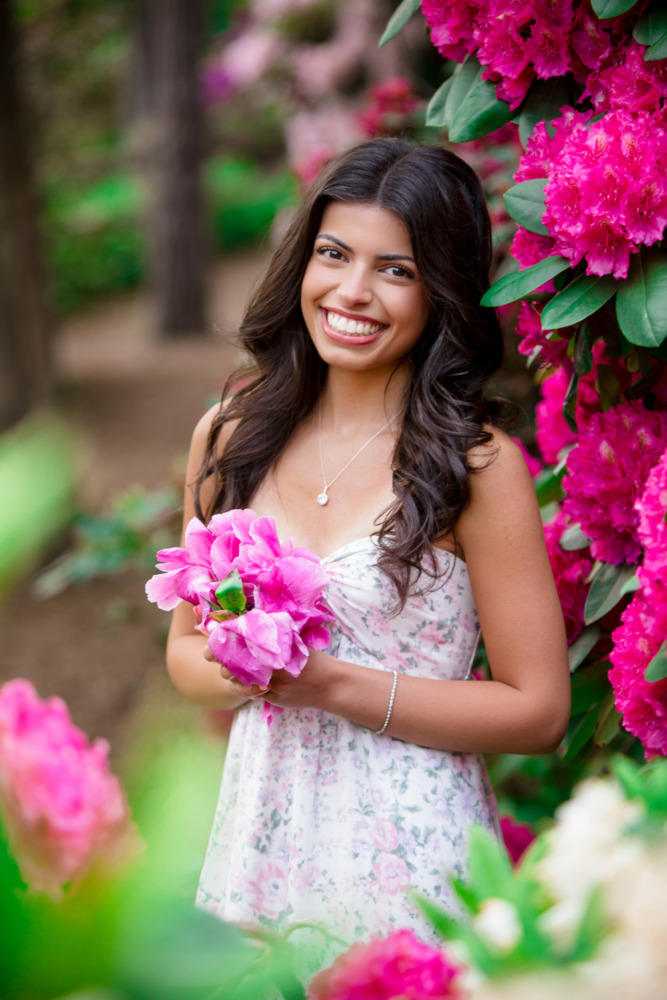 A high school senior girl with long dark hair, wearing a floral dress, smiles while holding bright pink flowers—capturing the perfect senior pictures with flowers beside a bush covered in vibrant pink blossoms outdoors.