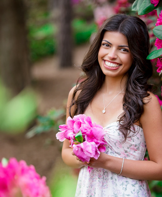 A high school senior girl in a floral dress stands outdoors, smiling and holding pink flowers—surrounded by vibrant pink blossoms and greenery. This scene captures the perfect vibe for senior pictures with flowers, set against a beautifully blurred natural background.