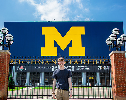 A high school senior guy walks outside the gates of Michigan Stadium during his Ann Arbor senior pictures session, with a large blue sign displaying a yellow "M" in the background, under a bright blue sky.