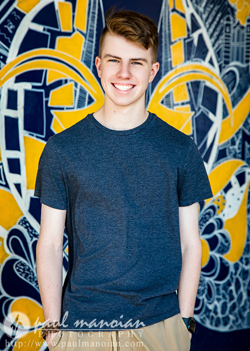 A high school senior guy with short light brown hair, wearing a dark gray t-shirt and khaki pants, smiles while standing in front of a vibrant mural with yellow, blue, and white abstract designs during his Ann Arbor senior pictures session.