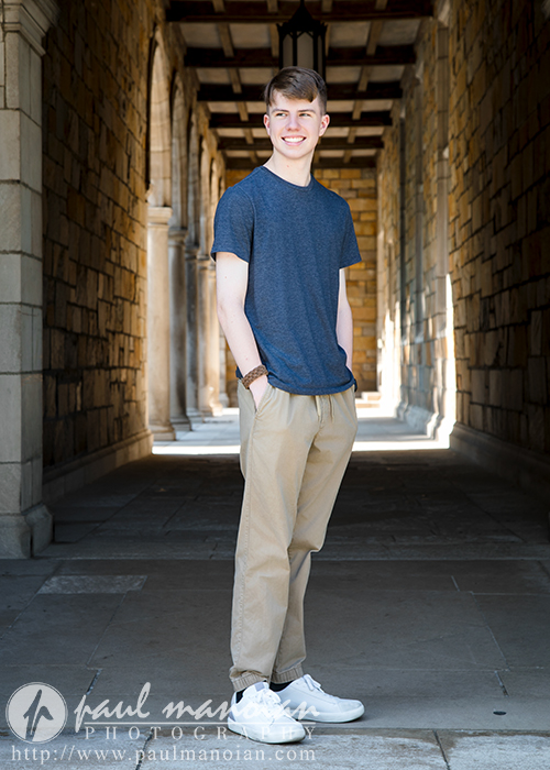 A high school senior boy in a blue t-shirt and khaki pants stands smiling in a sunlit stone walkway with archways during his Ann Arbor senior pictures session. The photographer’s watermark is visible at the bottom of the image.