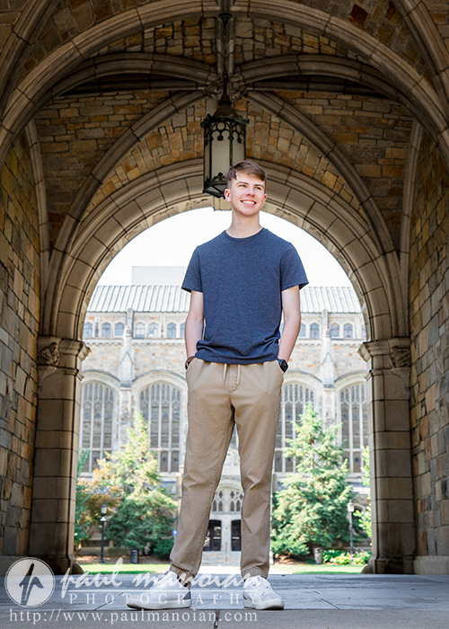 A high school senior in a blue t-shirt and khaki pants stands smiling under a large stone archway during his Ann Arbor senior pictures session, with a historic building and trees visible in the background on a sunny day.