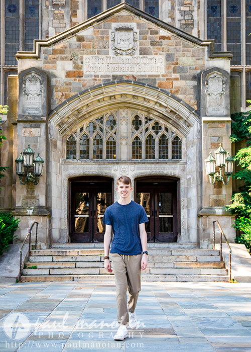 A high school senior boy in a blue t-shirt and khaki pants walks on a stone path during his Ann Arbor senior pictures session, in front of a large, ornate Gothic building with arched windows, steps, and greenery on the sides.