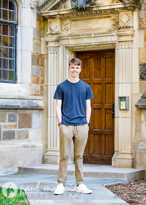 A high school senior guy stands smiling in front of a historic building with a wooden door, stone steps, and ornate stonework during his Ann Arbor senior pictures session. He wears a navy t-shirt, khaki pants, and white sneakers.