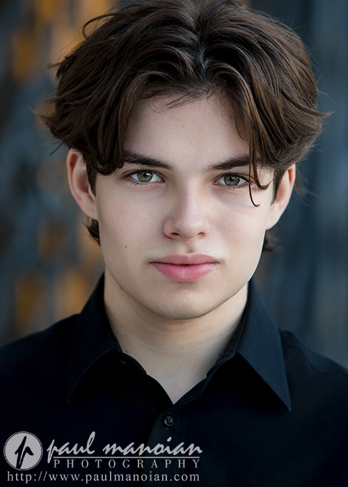 A young actor with fair skin, dark brown hair, and green eyes looks directly at the camera in this Auburn Hills actor headshots session, wearing a black collared shirt against a blurred background. The Paul Manoian Photography logo is visible at the bottom.