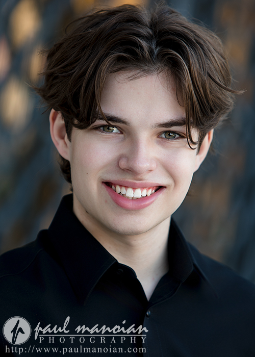 A young actor with medium-length brown hair and light eyes smiles at the camera, wearing a black collared shirt. The softly blurred background enhances this Auburn Hills actor headshots image, watermarked for Paul Manoian Photography.