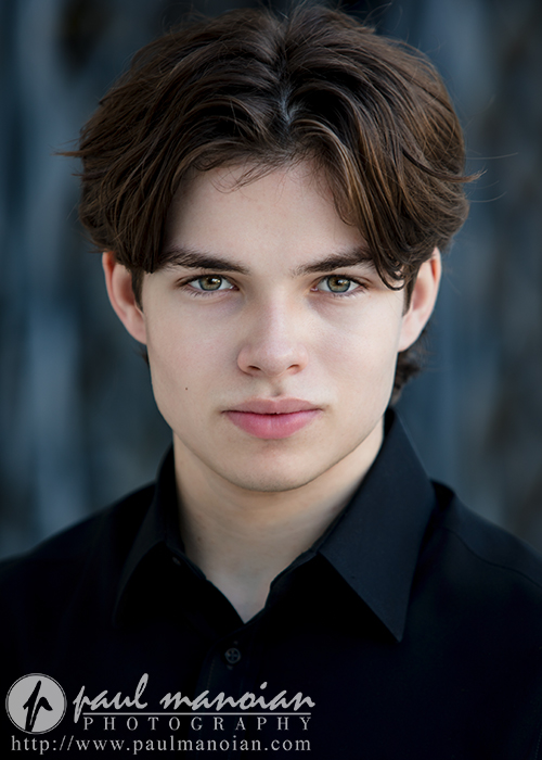 A young actor with brown hair and light eyes wearing a black collared shirt looks directly at the camera—an ideal example of Auburn Hills actor headshots. The background is softly blurred, with Paul Manoian Photography’s logo and website below.
