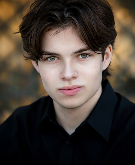 A young actor with fair skin, wavy brown hair, and green eyes, wearing a black shirt, looks directly at the camera with a neutral expression—an ideal look for Auburn Hills actor headshots. The background is softly blurred with warm tones.