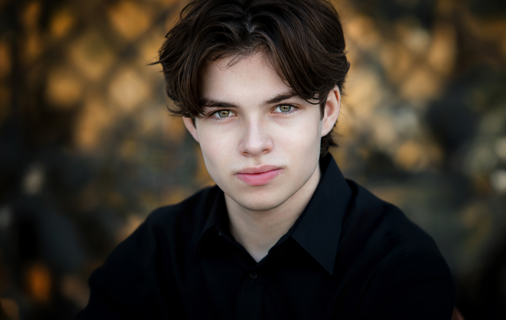 A young actor with fair skin, wavy brown hair, and green eyes, wearing a black shirt, looks directly at the camera with a neutral expression—an ideal look for Auburn Hills actor headshots. The background is softly blurred with warm tones.