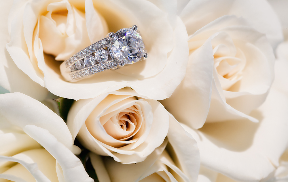 A sparkling diamond engagement ring with a band encrusted with smaller diamonds rests on a bouquet of ivory roses.