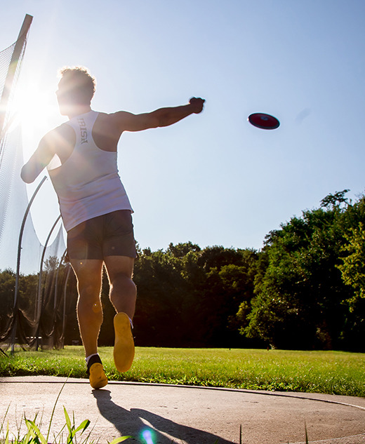 A high school senior throws a discus during a sunny outdoor practice, with the sun shining brightly behind them and trees in the background. The athlete is in mid-motion inside a throwing circle for his senior pictures session.