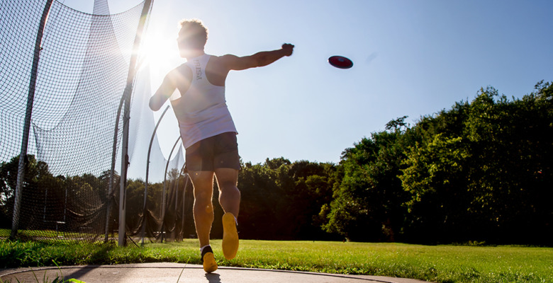 A high school senior throws a discus during a sunny outdoor practice, with the sun shining brightly behind them and trees in the background. The athlete is in mid-motion inside a throwing circle for his senior pictures session.