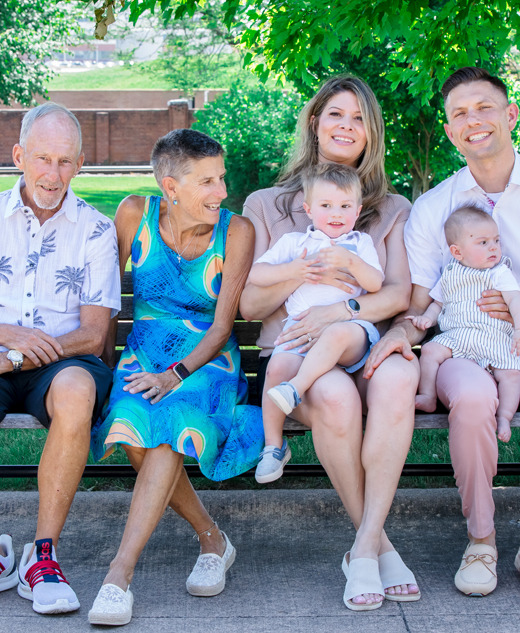 Three adults, including an older man and woman, sit on a park bench with a younger couple holding two babies for a Farmington Hills family portraits session with family photographer, Paul Manoian. Lush green trees and grass frame this sunny day—perfect for Farmington Hills family portraits.
