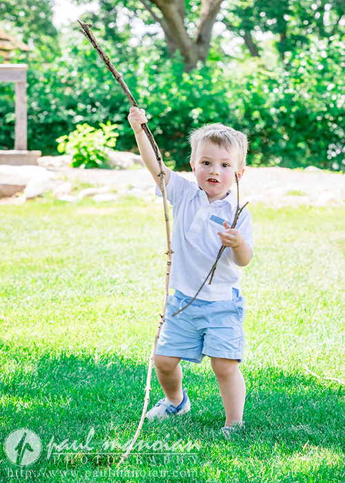 A young boy in a white shirt and blue shorts stands on green grass outdoors, holding two long sticks and smiling. Trees and sunlight fill the background.