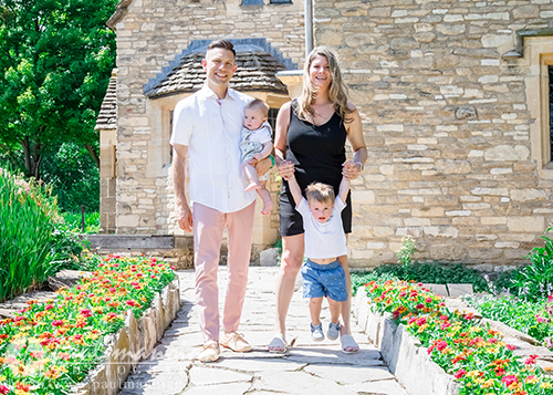 A smiling family of four poses on a stone path lined with red flowers, in front of a stone house for a family portrait session. The mother holds a toddler's arms, lifting him mid-air, while the father holds a baby. It is a bright, sunny day.