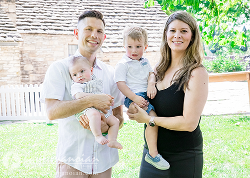 A smiling couple poses outdoors with their two young children for a family portrait session; the man holds a baby while the woman holds a toddler. They stand on grass in front of a wooden house and white picket fence.