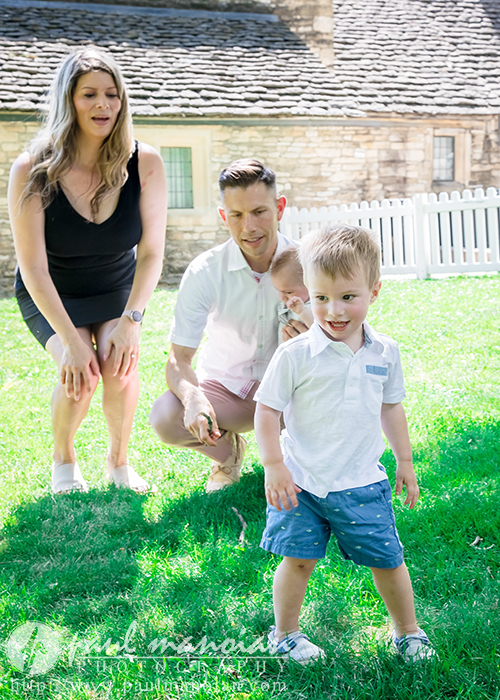 A smiling young boy in a white shirt and blue shorts walks on grass while a woman, man, and baby kneel and watch him during a family portrait session in a sunny backyard with a white fence and stone house in the background.
