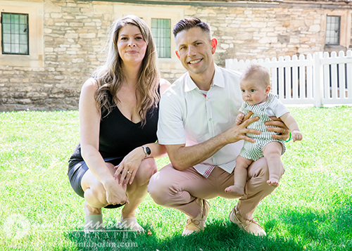A woman, a man, and a baby pose together on grass in front of a stone building and white picket fence for a family portrait session. The woman is kneeling, the man is squatting, and he holds the baby on his knee.