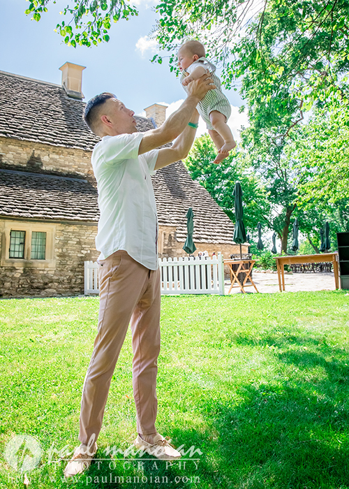 A man in a white shirt and pink pants lifts a smiling baby into the air outside, with a stone cottage, green grass, and trees in the background on a sunny day for a family portrait session.
