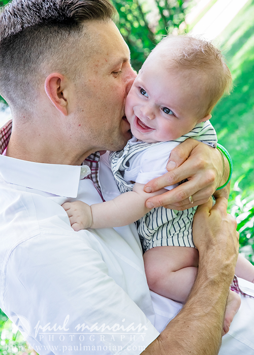 A man lovingly kisses a smiling baby on the cheek while holding the child in his arms outdoors during a family portrait session, surrounded by greenery. The baby looks happy and content, wearing a striped outfit.