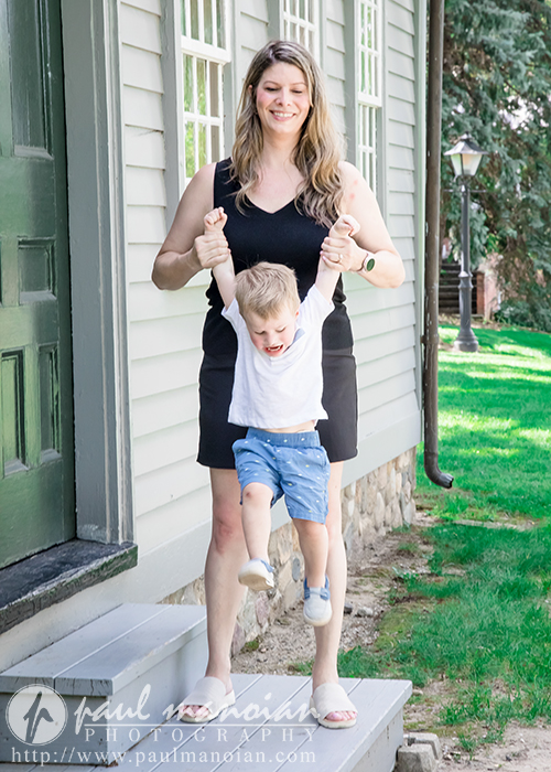 A smiling woman in a black dress holds a young boy's hands, lifting him as they step off a porch during a family portrait session. The boy, in a white shirt and blue shorts, swings joyfully in front of a gray house with green steps and a lawn.