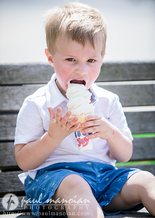 A young boy in a white shirt and blue shorts sits on a bench, holding and licking a large vanilla ice cream cone, enjoying a sunny day during a family portrait session.