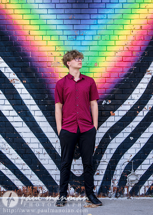 A high school senior in a red patterned shirt and black pants stands in front of a colorful mural with a rainbow heart and black-and-white striped wings on a brick wall. He looks to the side with his hands in his pockets.