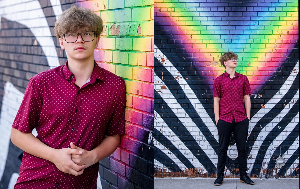A young person with glasses and wavy hair, wearing a red polka dot shirt and black pants, poses in front of a colorful mural with rainbow and black-and-white patterns on a brick wall.