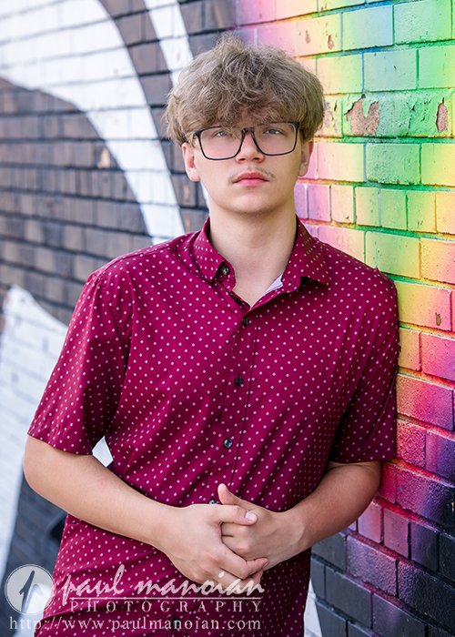 A high school senior with curly, light brown hair and glasses wears a maroon, polka dot shirt while standing against a colorful, graffiti-covered brick wall.