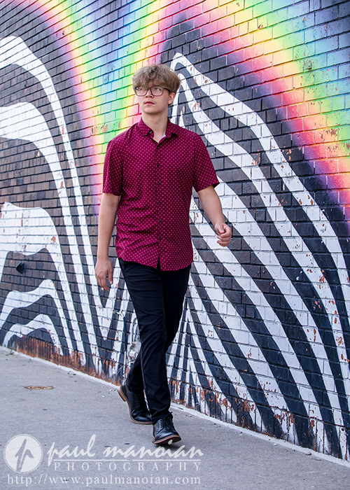 A high school senior with blond hair, wearing glasses, a red patterned shirt, and black pants walks past a mural featuring black and white zebra stripes and a rainbow on a brick wall.