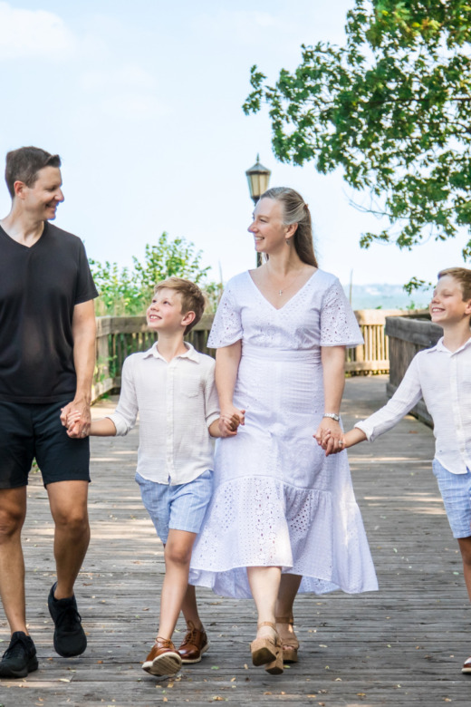 A family of four, two adults and two young boys, walk hand in hand on a wooden boardwalk outdoors, smiling and enjoying a sunny day surrounded by greenery and lanterns—captured by a Grosse Pointe family portraits photographer.