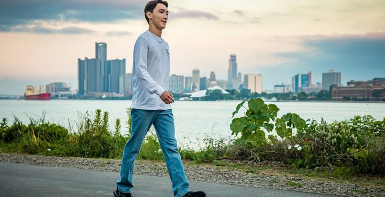 A high school senior boy in jeans and a white long-sleeve shirt walks on a riverside path with a city skyline and water in the background during a cloudy sunset, capturing moments from his Livonia senior pictures session.