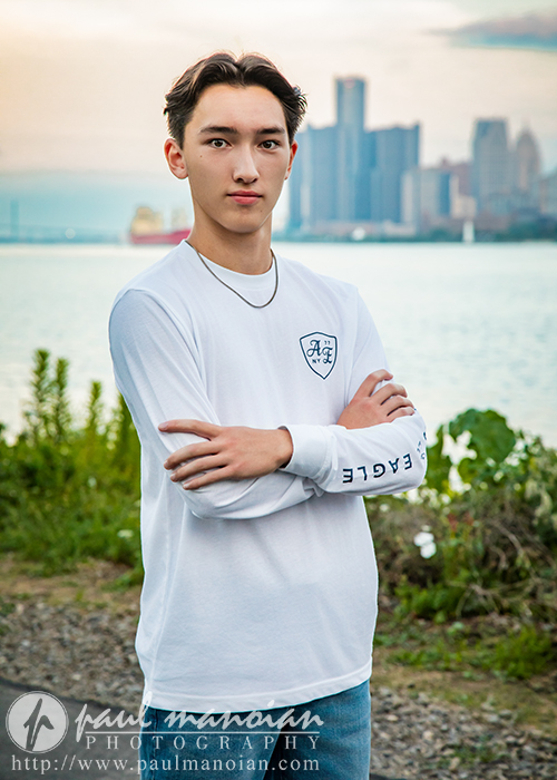 A high school senior boy with short brown hair stands outdoors by a river, crossing his arms and looking at the camera during his Livonia senior pictures session. He wears a white "EAGLE" shirt, with a city skyline in the background.