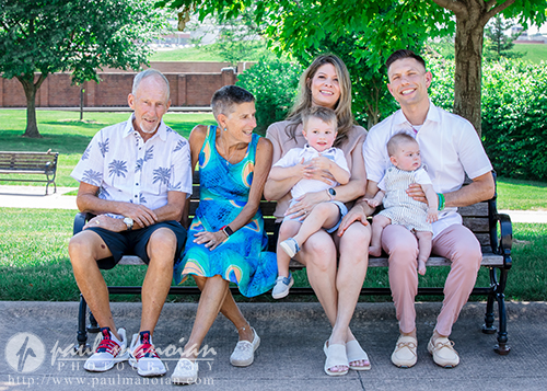 Four adults and two babies sit smiling on a park bench under a tree on a sunny day for a family portrait session. The adults appear to be two older people on the left and two younger on the right, holding the babies. Green grass and benches are visible in the background.