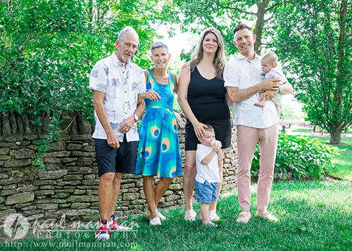 Three adults and two children stand barefoot on grass in front of a stone wall and trees, posing for a family photo on a sunny day. One adult holds a baby, and a child stands near an adult’s leg.
