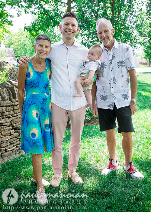 Three adults and a baby stand together outside on grass, smiling at the camera. Two older adults stand on either side of a younger man holding the baby during a family portrait session. Trees and a stone wall are in the background.