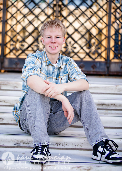 A high school senior guy with short blond hair, wearing a plaid shirt and light gray jeans, sits smiling on outdoor white steps in front of an ornate metal gate during his Novi senior pictures session.