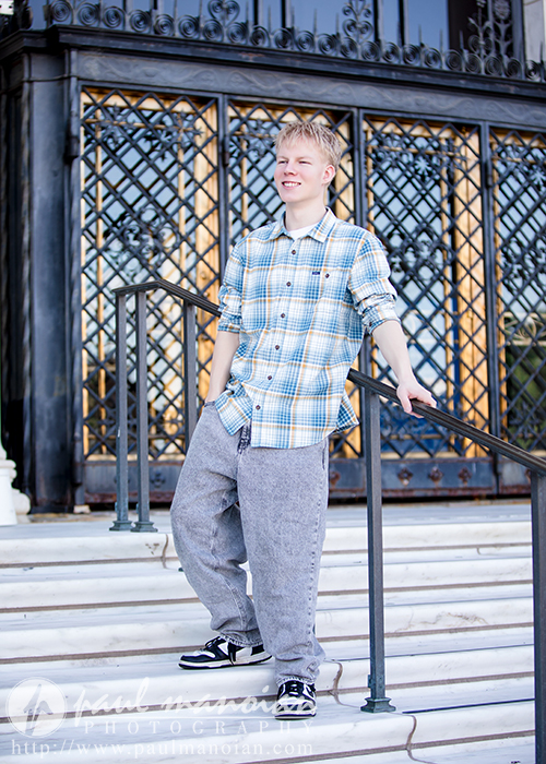A smiling high school senior guy in a blue plaid shirt and gray pants stands on marble steps, holding a railing, with ornate black and gold doors in the background during his Novi senior pictures session.
