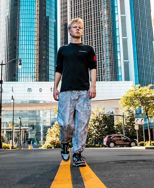 A high school senior guy walks down the center of a city street between tall buildings, with the Renaissance Center skyscrapers and a bright blue sky in the background, capturing a memorable moment from his Novi senior pictures session.